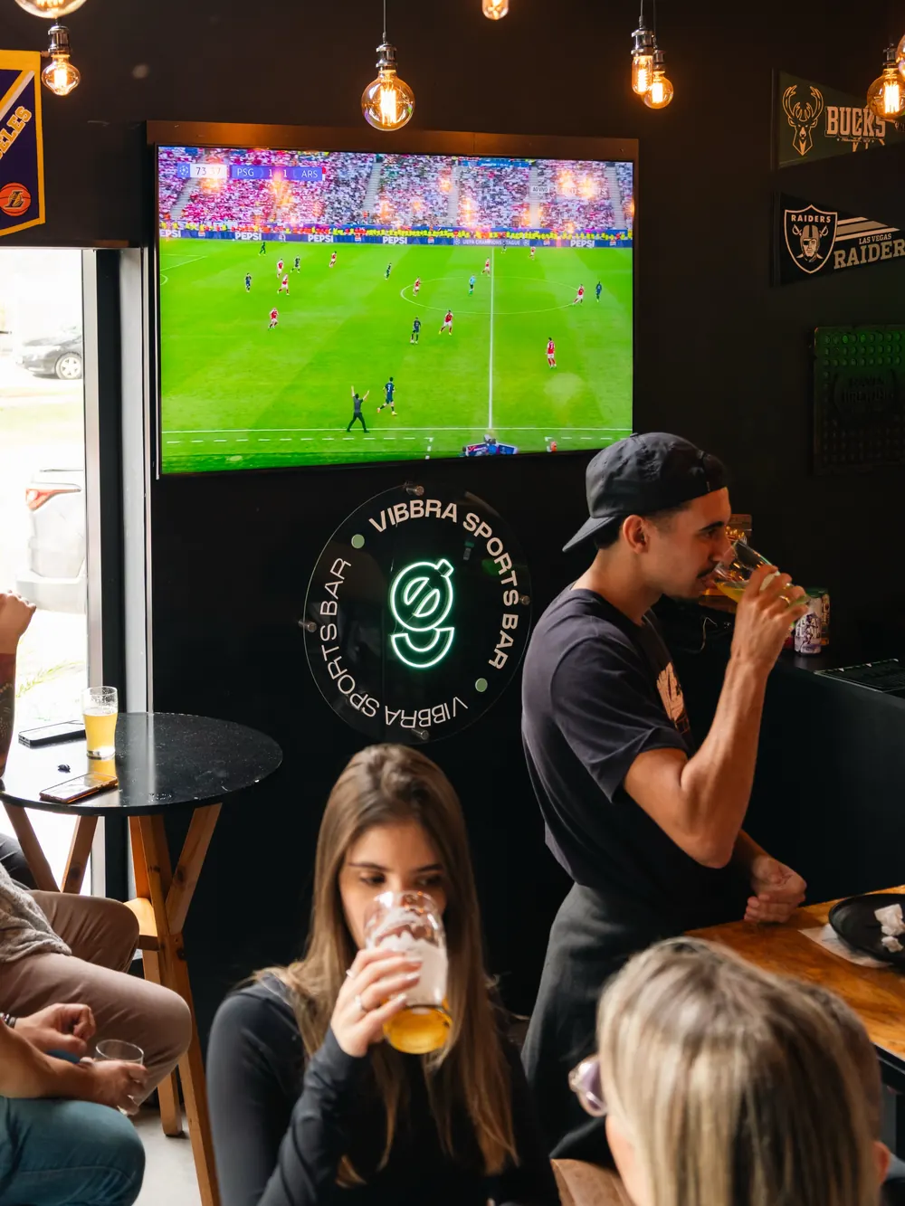 Clientes bebendo chope em frente ao telão com uma partida de futebol